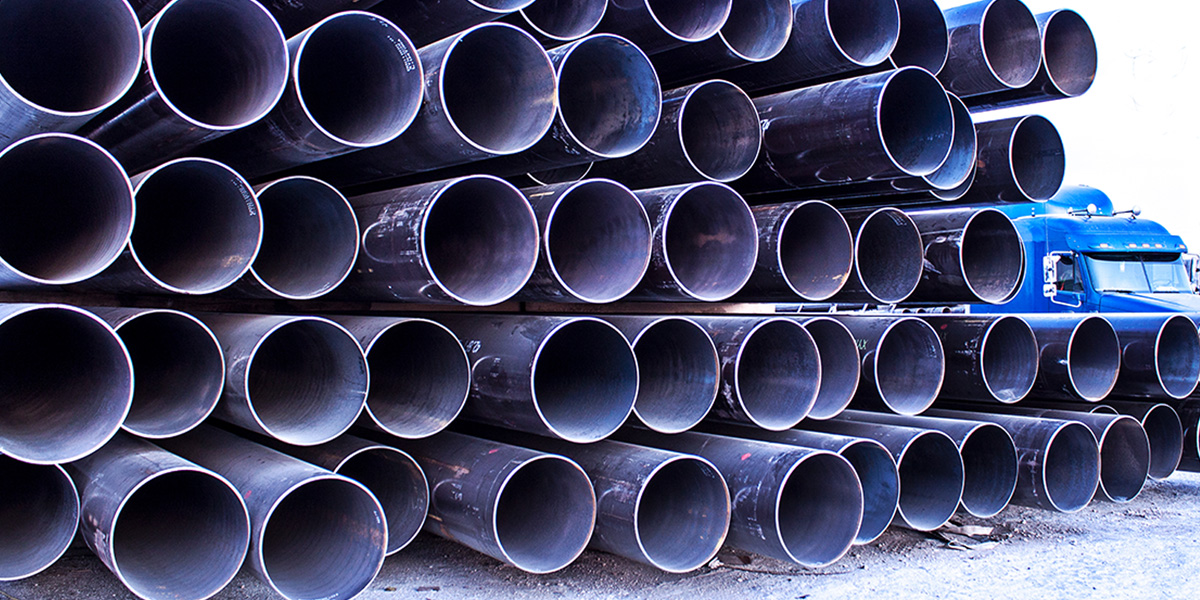 Stacks of large diameter welded carbon steel pipe arranged next to a truck in a pipe yard.